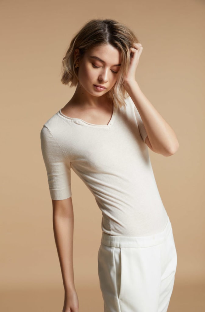 A lookbook fashion image featuring a female model wearing a creamy top and white colour pants, resting her hand on her forehead in front of a beige background. Photographed by I Heart Studios.