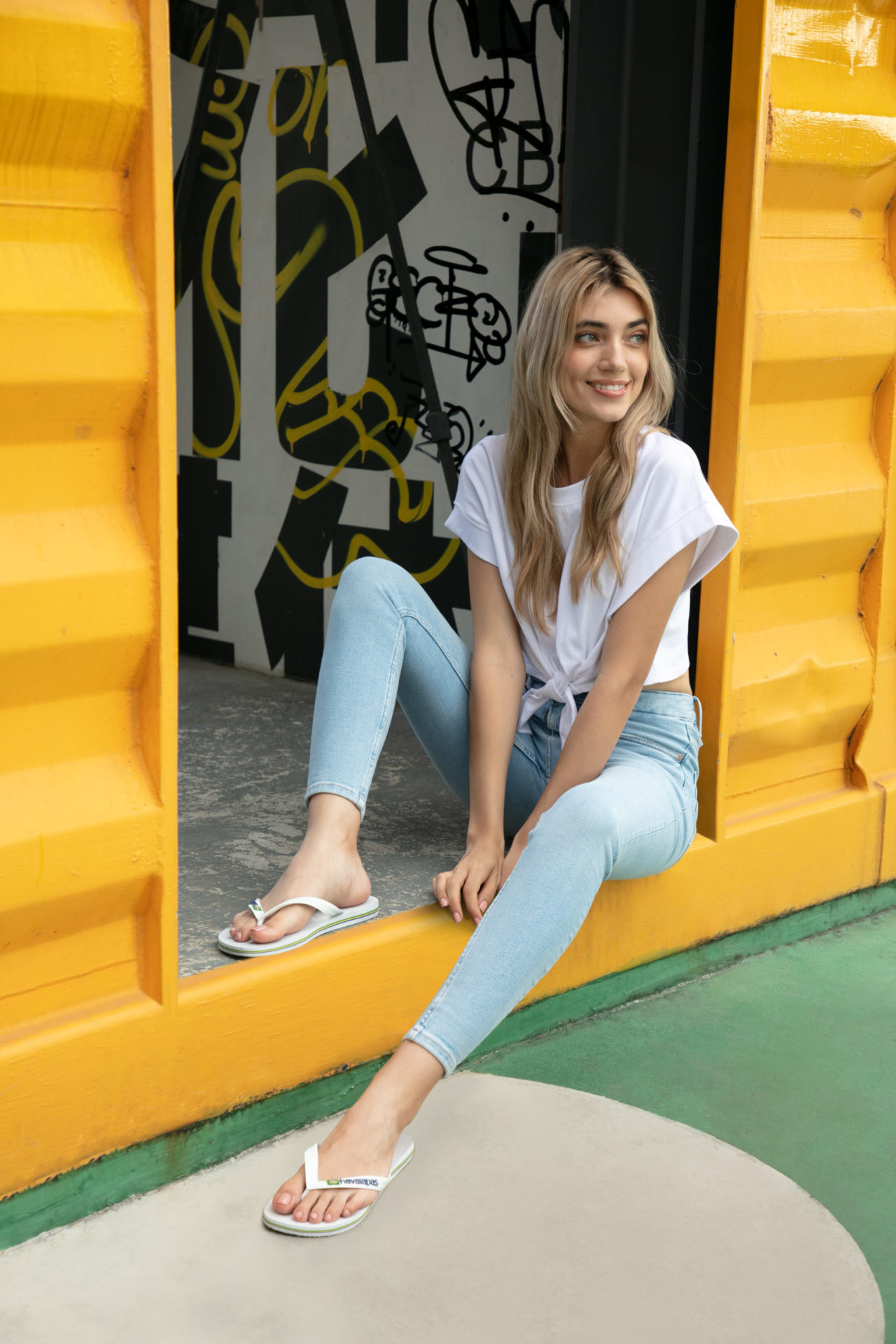 An image depicting a joyful woman wearing a white top and blue jeans, sitting on the step of a yellow shipping container with a vibrant smile. This creative promotional image was captured by I Heart Studios.
