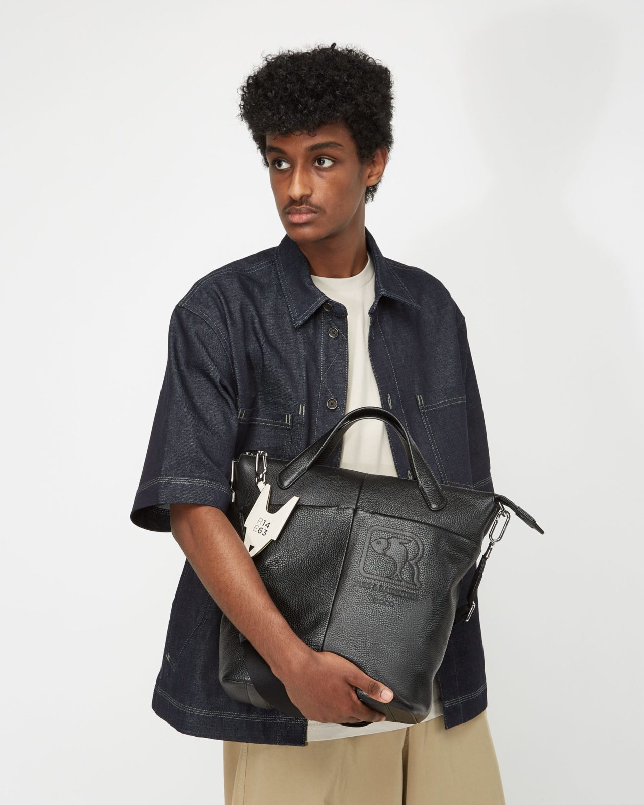 A lookbook fashion image featuring a man wearing a fashionable outfit holding a bag with his hand in front of a white background. Photographed by I Heart Studios.