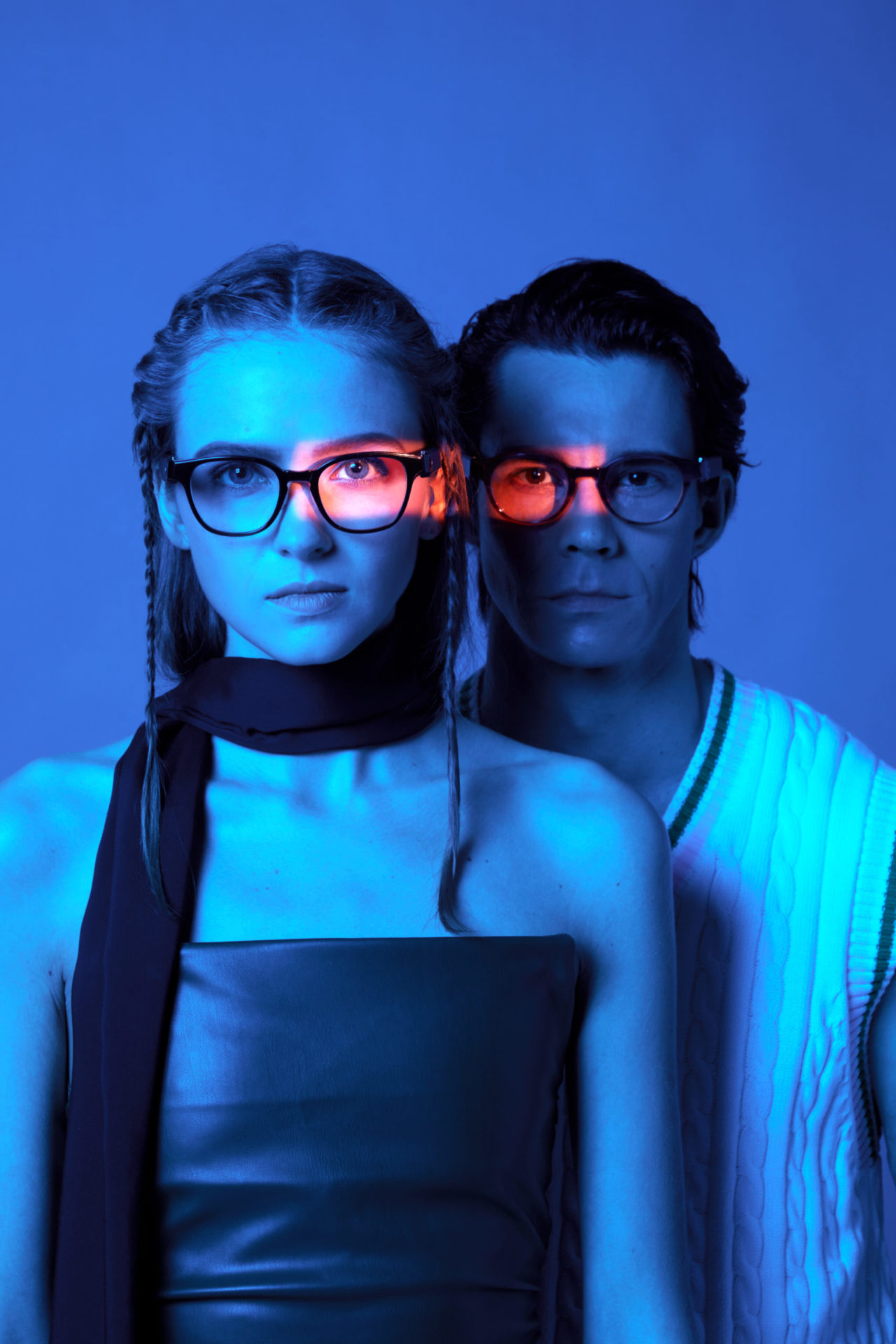 A man and a woman wearing eyeglasses. The background features a blue background with a neon red lighting effecton their eyeglasses. This creative promotional shoot was captured by I Heart Studios.