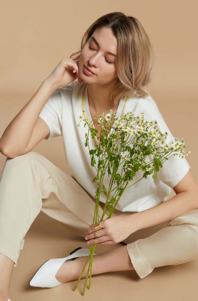 A lookbook fashion image featuring a female model wearing a white top and creamy colour pants, holding a bouquet with her hand sitting in front of a beige background. Photographed by I Heart Studios.