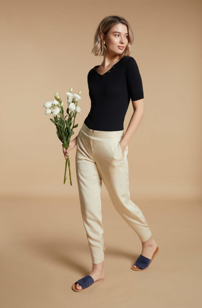 A lookbook fashion image featuring a female model wearing a black top and a white pant, holding a bouquet with her hand in front of a beige background. Photographed by I Heart Studios.