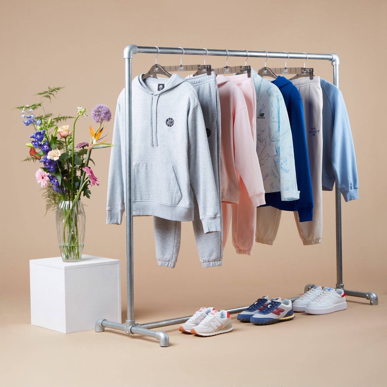 An image showcasing a clothing rack with sports clothes and three pairs of sneakers. Adjacent to the rack, there is a white cube with a vase containing a flower. Creative promotional image captured by I Heart Studios.