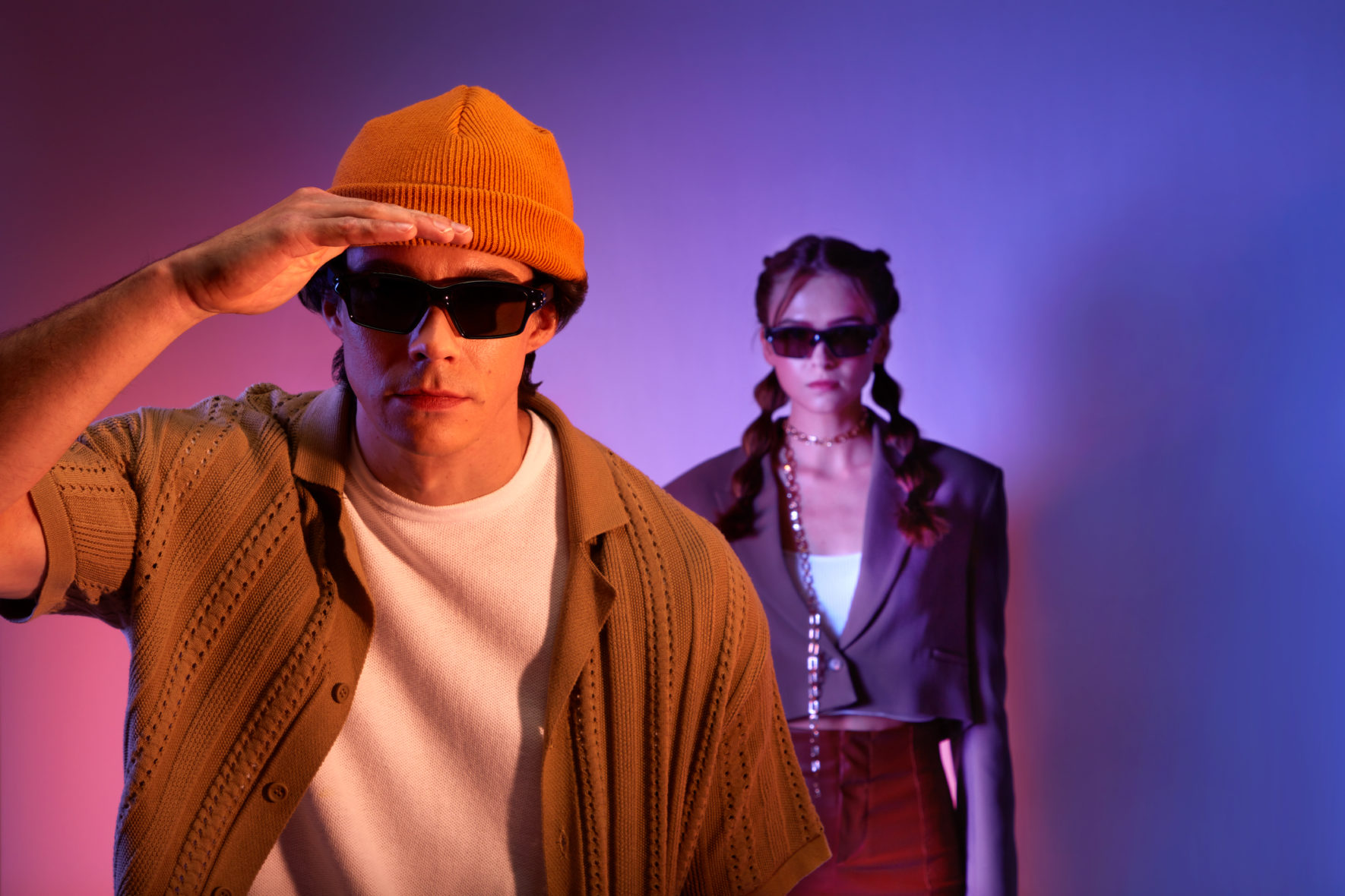 A man and a woman wearing sunglasses, with the man looking into the distance and placing his hand on his forehead. The background features pink and purple colors with a lighting effect. This creative promotional shoot was captured by I Heart Studios.