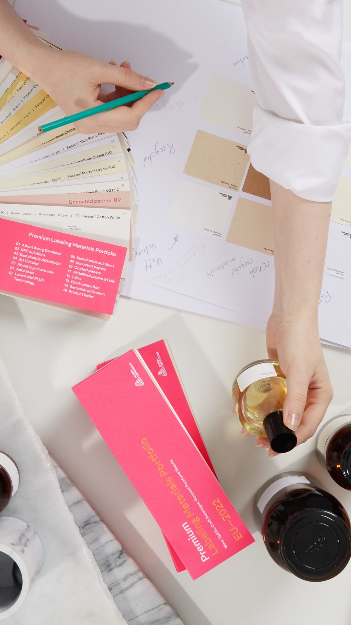 A woman carefully applying a sticker to a bottle package on her neatly organized working table, surrounded by a color chart and a stack of brown packaging materials. Photographed by I Heart Studios.