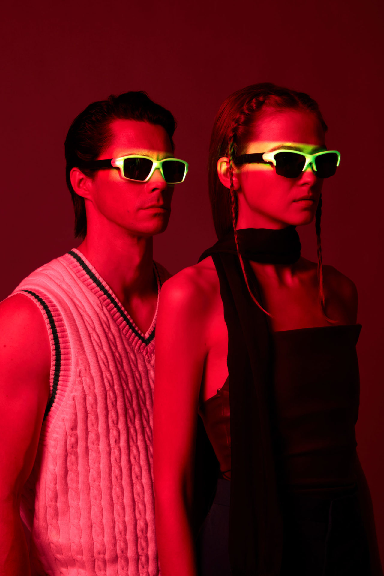 A man and a woman wearing sunglasses, with the man and women both looking into the distance. The background features a red color with a neon green lighting effect on the sunglasses part to highlight the product. This creative promotional shoot was captured by I Heart Studios.